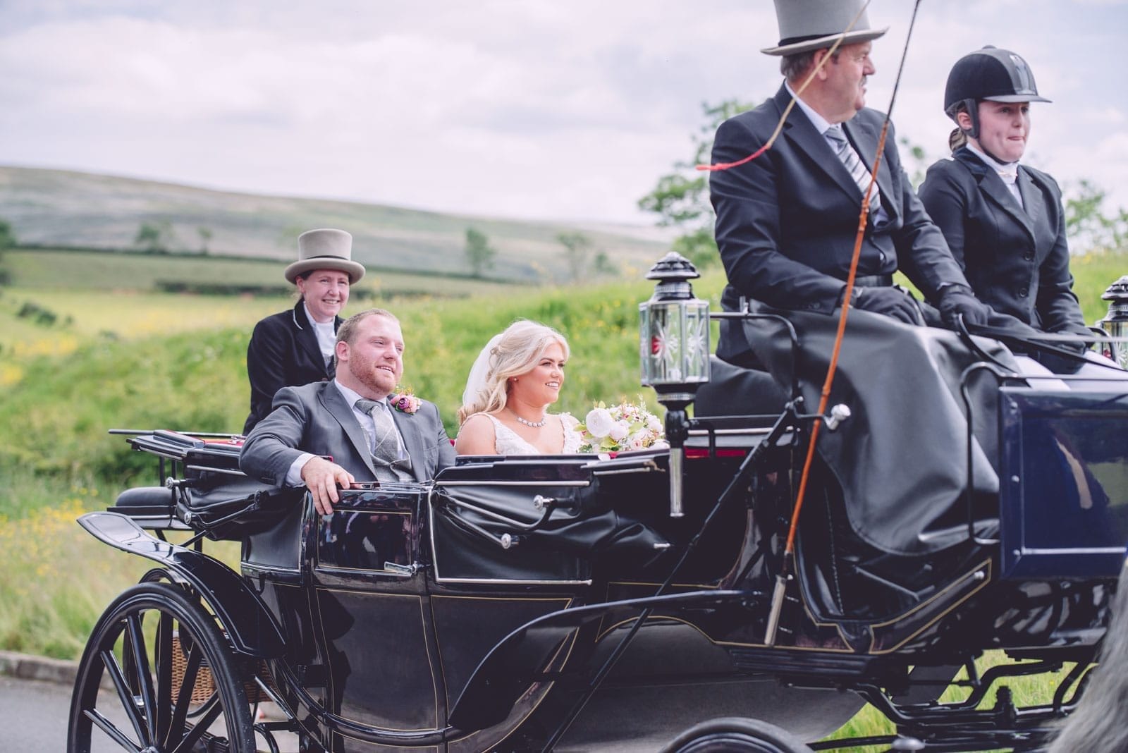 A couple in wedding attire sit in a horse-drawn carriage, captured beautifully by a wedding photographer with coachmen in top hats and a rural background. northern ireland wedding photographer