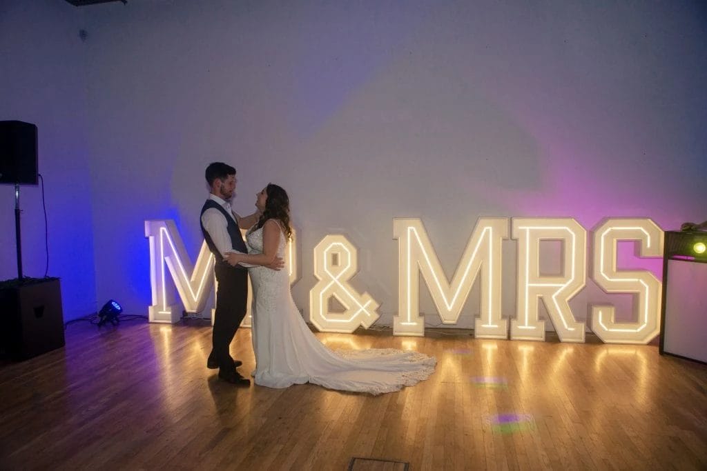 A bride and groom dance in front of large illuminated letters spelling "MR & MRS" on a wooden floor in a dimly lit room, perfectly captured by their Ballymoney wedding photographer. northern ireland wedding photographer